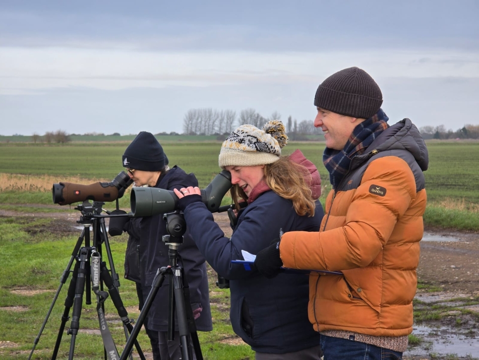 Three people, two with telescopes, one with a clipboard monitoring swans out of view.